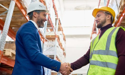 Recruitment Deal Waist up portrait of handsome mature businessman shaking hands with worker wearing hardhat standing against warehouse shelves in background